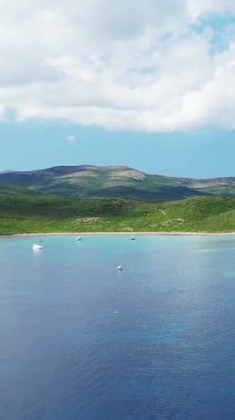 Finocchiarola körfezinin turkuaz suyu, yeşil tepeler ve parçalı bulutlu bir gökyüzünün altında demirli yelkenlileri olan manzarası. Huzurlu kıyı manzarası seyahat ve doğa kavramları için ideal..
