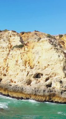 Portekiz 'in Lagos kenti yakınlarındaki Algarve kıyısındaki turkuaz suların üzerinde açık mavi gökyüzünün altında dramatik altın uçurumlar yükseliyor. Doğal kaya oluşumları ve canlı renkler çarpıcı bir kıyı manzarası yaratır..