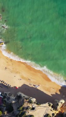 Portekiz 'in Albufeira yakınlarındaki Algarve kıyısında turkuaz dalgalar ve kayalık uçurumlarla kumlu bir sahilin insansız hava aracı görüntüleri. Güneş banyosu ve şemsiyeler altın kumda görünür..