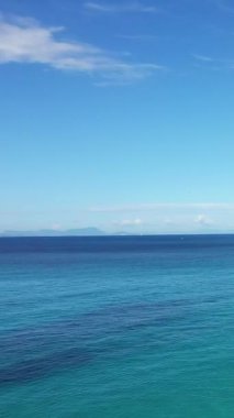 Berrak turkuaz suyu ve açık mavi gökyüzü altındaki uzak dağlarla sakin deniz manzarası, Karavostasi, Yunanistan.