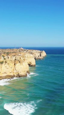 Portekiz 'in Lagos yakınlarındaki Algarve kıyı şeridi boyunca turkuaz okyanus dalgalarının üzerinde, açık mavi bir gökyüzünün altında dramatik altın uçurumlar yükseliyor..
