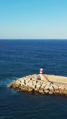 Portekiz 'in Sagres kenti yakınlarındaki Algarve kıyısı boyunca uzanan mavi denize uzanan kayalık bir dalgakıran üzerinde kırmızı ve beyaz bir deniz feneri duruyor. Açık gökyüzü ve sakin okyanus sakin bir kıyı manzarası yaratır..