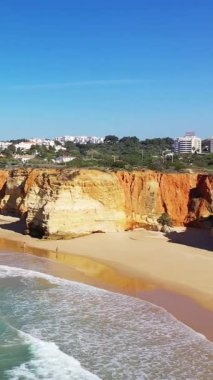 Portekiz 'in Albufeira yakınlarındaki Algarve kıyısı boyunca dramatik uçurumlar ve turkuaz dalgaların olduğu kumlu bir sahil manzarası. Modern binalar arka planda berrak mavi gökyüzünün altında görülebilir..