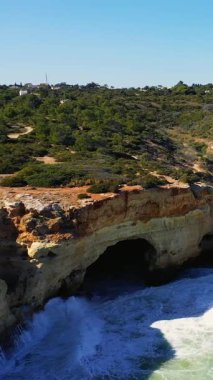 Portekiz 'deki Algar de Benagil' de okyanusun yukarısındaki dramatik deniz kayalıkları ve yemyeşil yeşil, dalgalar kayalık bir mağaraya çarpıyor. Açık mavi gökyüzünün altındaki parlak doğal manzara.