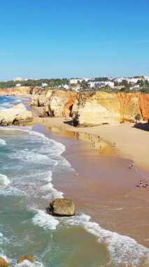 Portekiz 'in Albufeira yakınlarındaki Algarve kıyısı boyunca dramatik uçurumlar ve turkuaz dalgalarla kumlu sahilin manzarası. İnsanlar açık mavi gökyüzünün altında kıyı boyunca yürür..