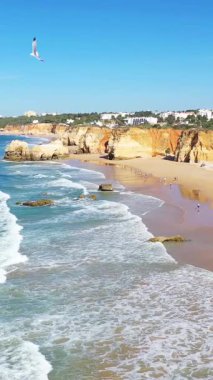 Albufeira yakınlarındaki Algarve sahilinin manzarasında açık mavi gökyüzü altında altın kayalıklar, kumlu plajlar ve turkuaz dalgalar var..