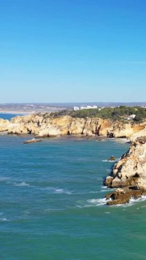 Portekiz 'in Albufeira kenti yakınlarındaki Algarve kıyısındaki engebeli kayalıkların ve turkuaz suların berrak mavi gökyüzünün altında manzarası..