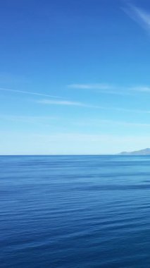 Berrak mavi suyu olan sakin bir deniz manzarası ve parlak bir gökyüzü altında uzak bir kıyı şeridi, Palinuro, İtalya.