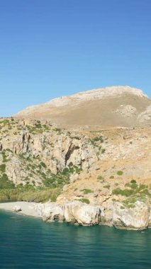 Girit, Preveli 'deki kayalık kıyı şeridi ve kumlu sahil manzarası berrak mavi sular ve engebeli tepeler parlak bir gökyüzü altında..