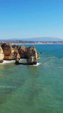 Portekiz 'in Lagos yakınlarındaki Algarve kıyısı boyunca açık mavi gökyüzünün altında, turkuaz suların üzerinde dramatik kireçtaşı kayalıkları yükseliyor..