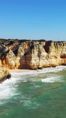 Portekiz 'in Lagos kenti yakınlarındaki Algarve kıyısında açık mavi bir gökyüzünün altında kumlu bir plaj ve turkuaz dalgalar üzerinde dramatik kireçtaşı uçurumları yükseliyor..
