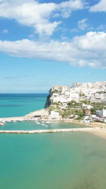 Peschici 'nin turkuaz suların üzerindeki bir uçuruma tünemiş beyaz binalar ve parlak bir gökyüzünün altındaki küçük bir marina manzarası. Resimli Adriyatik kıyı şeridi güneşli bir günde görünür.