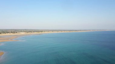 Geniş Grande Plage de Barneville 'in sakin mavi denizi ve açık gökyüzünün altındaki kumlu kıyı şeridi. Uzak kasaba ve doğal güzelliklerle kıyı manzarası.