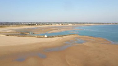 Geniş kumsal manzarası ve Grande Plage de Barneville, Normandiya, Fransa 'daki sakin mavi sular. Sahnede gelgit havuzları, dalgalar ve açık gökyüzü altında uzak bir kasaba yer alıyor..