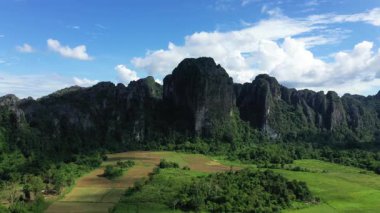 Vang Vieng, Laos 'u çevreleyen kırsaldaki yemyeşil tarlaların ve dramatik kireçtaşı uçurumların havadan görünüşü, dağınık mavi bulutlarla dolu bir gökyüzünün altında..