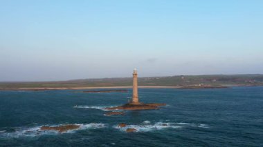 Fransa 'nın Normandiya, Cap de la Hague yakınlarındaki bir deniz kıyısındaki kayalık bir adada uzun bir deniz feneri duruyor. Sahil şeridi ve açık deniz berrak mavi gökyüzünün altında görülebilir..