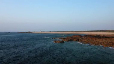 Fransa 'nın başkenti Cap de la Hague' deki engebeli kıyı şeridi ve kumlu sahilin geniş manzarası berrak mavi suları ve sakin bir gökyüzünün altındaki uzak deniz feneriyle.