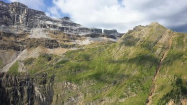 Fransız Pireneleri 'ndeki Gavarnie Sirki' nin dramatik kayalık yamaçları ve yeşil yamaçları parçalı bulutlu bir gökyüzünün altında..