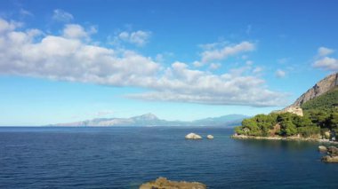 Scenic view of the Maratea coastline with clear blue sea, rocky shore, and lush green hills under a bright sky with scattered clouds.