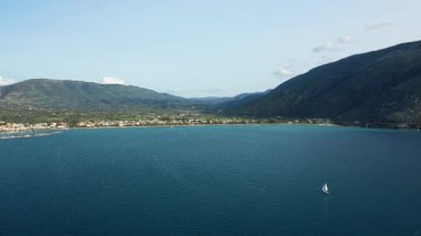 A lone sailboat glides across tranquil blue waters with the scenic coastline and mountains of Plataria, Greece in the background under a clear sky.