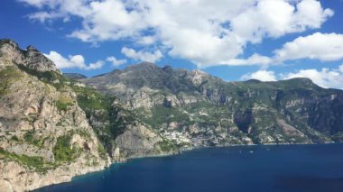 İtalya 'nın Positano yakınlarındaki Amalfi kıyısındaki engebeli kayalıkların ve turkuaz denizin parlak mavi gökyüzünün altında dağınık bulutlarla nefes kesen görüntüsü..