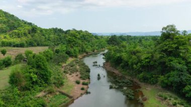 Laos kırsalındaki yoğun yeşil bitki örtüsü ve orman tepeleriyle çevrili dolambaçlı bir nehrin havadan görünüşü. Parçalı bulutlu bir gökyüzü altında sakin doğal manzara.