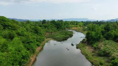 Laos kırsalında yoğun yeşil bitki örtüsü ve uzak dağlarla çevrili dolambaçlı bir nehrin havadan görünüşü. Parlak gökyüzünün altındaki sakin doğal manzara.