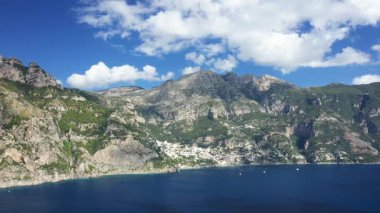 Panoramic view of Positano with dramatic cliffs, lush green mountains, and the blue waters of the Amalfi Coast under a partly cloudy sky.