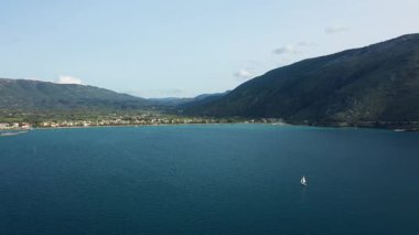 Tek bir yelkenli Plataria Körfezi 'nin sakin mavi sularında süzülüyor. Dağlar ve arka planda berrak bir gökyüzünün altında bir kıyı köyü var..