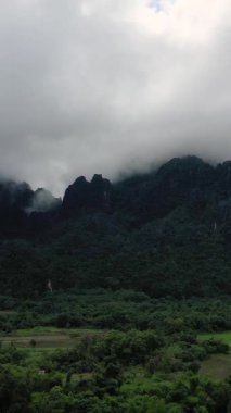 Yoğun yeşil ormanın ve Laos 'un Vang Vieng yakınlarındaki tarlaların üzerinde sisle kaplanmış dramatik kireçtaşı dağları. Bulutlu gökyüzünün altında yemyeşil bir tropikal arazi.