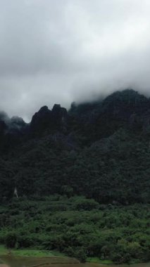 Yoğun yeşil orman, Laos, Vang Vieng yakınlarında sisle kaplanmış dramatik kireçtaşı dağlarını kaplıyor. Bulutlu gökyüzünün altında yemyeşil bir tropikal arazi.