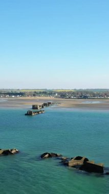 Arkaplanda açık mavi gökyüzü ve kıyı köyü bulunan Gold Beach, Asnelles, Normandiya, Fransa 'ya uzanan tarihi yapay liman kalıntılarının hava manzarası.