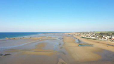 Geniş kumlu kıyı şeridi ve Fransa 'nın Asnelles kentindeki Gold Beach' teki sakin mavi denizin kenarları boyunca görünen küçük bir kıyı köyü. Açık gökyüzü ve sakin atmosfer manzaralı kıyı şeridini vurguluyor.