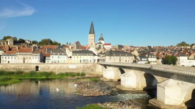 La Charite sur Loire 'ın ikonik taş köprüsü, geleneksel evleri ve açık mavi gökyüzünün altındaki kilise manzarası. Loire Nehri, tarihi mimarinin altından nazikçe akar..