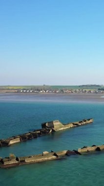 Fransa 'nın Asnelles kentindeki Gold Beach sahil şeridini geniş açıdan görebiliyoruz. Durgun turkuaz sular, kumlu kıyılar ve berrak mavi gökyüzü altında yapay bir liman yapısının görünür bir bölümü var..