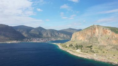 Yunanistan 'ın Monemvasia kıyısındaki engebeli uçurumlar ve derin mavi deniz manzaralı, parçalı bulutlu bir gökyüzü altında. Manzarada dramatik kaya oluşumları ve kıyı şeridinde uzak bir kasaba yer alıyor..