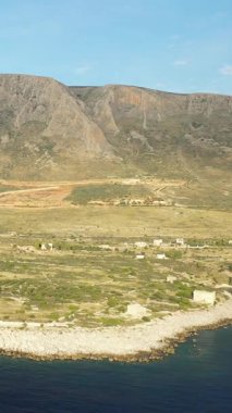 Yunanistan 'ın Cap Tenare kentinin kayalık kıyı şeridi ve dalgalı tepelerinin geniş manzarası, dağınık küçük binalar ve parçalı bulutlu bir gökyüzü altında sakin mavi deniz.