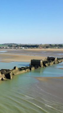 Alçak gelgitte Gold Beach 'in geniş manzarası, kumlu kıyıları, sakin suyu ve Asnelles, Normandiya, Fransa' daki uzak köy binalarını gösteriyor..