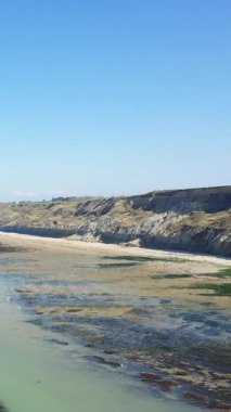 Port en Bessin, Normandiya, Fransa 'da açık mavi gökyüzünün altında engebeli kayalıklar ve kumlu plaj manzarası. Açık kıyı şeridi ve sakin atmosferi olan doğal kıyı manzarası.