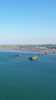 Asnelles, Normandiya yakınlarındaki Gold Beach açıklarındaki mavi sulardaki yapay liman kalıntılarının hava görüntüsü, arka planda açık bir gökyüzü ve kıyı manzarası var..
