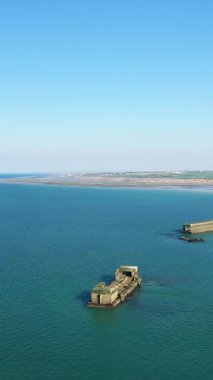 Gold Beach, Asnelles, Normandiya, Fransa 'daki sakin mavi sularda bulunan yapay bir limandan beton yapıların hava görüntüsü. Kıyı şeridi ve uzak kırsal bölge berrak bir gökyüzünün altında görülebilir..