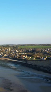 Arromanches-les-Bains 'in kıyı köyü, kumlu plajı ve açık mavi gökyüzü altında çevreleyen kırsal manzarası. Sahne Fransa, Normandiya 'nın sakin atmosferini yakalıyor..