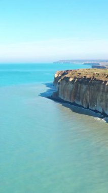 Dik tebeşir kayalıkları, açık mavi gökyüzünün altında Normandiya kıyısı boyunca turkuaz denizin üzerinde yükselir. Doğal manzara, ikonik kıyı manzarasını vurgular..