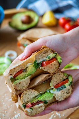 Avokado, mayonez, salatalık, marul ve soğanla ikiye bölünmüş içi doldurulmuş simit sandviçi tutan eller. Arka planda tütsülenmiş somon simidi. Glütensiz sağlıklı kahvaltı, canlı eğlenceli renkler.