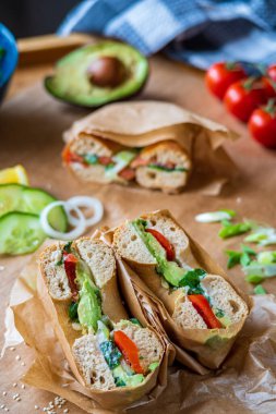Avokado, mayonez, salatalık, marul ve domatesle ikiye bölünmüş kızarmış simit sandviçi. Arka planda, kağıda sarılı somon simidi. Glütensiz sağlıklı kahvaltı, canlı eğlenceli renkler.