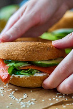Tütsülenmiş somon, krem peynir, salatalık ve marulla doldurulmuş simit sandviçini yakından çek. Sağlıklı bir kahvaltı için ev yapımı glütensiz kızarmış simit. Eğlenceli canlı renkler. Yan görünüm
