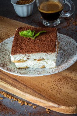 Arka planda bir fincan kahveyle servis edilen geleneksel tiramisu pastasından bir dilim. Otantik glütensiz İtalyan katmanlı tatlı, ladyfinger bisküvileri, mascarpone peynir kremi ve kakao tozu. Yan görünüm
