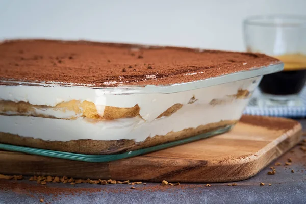 Traditional tiramisu cake in a glass pan on a wooden board served with a cup of coffee. Authentic gluten-free Italian layered dessert with ladyfinger biscuits, mascarpone cheese cream and cocoa powder