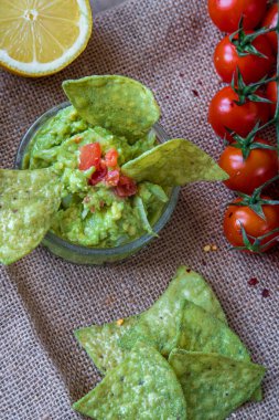 Taze ev yapımı avokado kasesi, yeşil, avokado aromalı, tortilla cipsi. Glütensiz geleneksel Meksika yemeği. Çeri domatesleri, avokado ve limon arka planda kahverengi, kırsal, kumaş. Üst görünüm