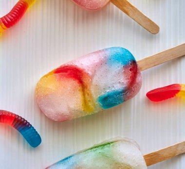 Beyaz bir tabakta renkli donmuş solucan şekerlemelerinin en üst görüntüsü, yaz aylarındaki çocuklar için harika ev yapımı buzlu şeker ve tahta çubuklu şeker solucanları.